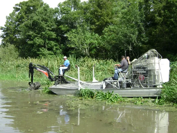 Faucheux airboat with mounted equipment working beside a wooded shallow-water bank